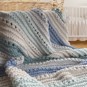 May include: A soft, striped knitted baby blanket in shades of blue, gray, and green. The blanket is folded and draped over a wicker basket.