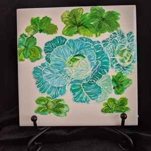 May include: A hand-painted ceramic tile featuring a green and blue cabbage with white veins. The cabbage is surrounded by smaller green leaves. The tile is on a black stand.