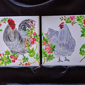 May include: Two ceramic tiles with hand-painted images of chickens. The tile on the left features a rooster with a red comb and a gray and white body. The tile on the right features a hen with a gray and white body. Both tiles feature pink flowers and green leaves.