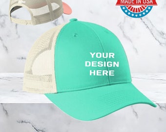 May include: Two blank trucker hats, one pink and one turquoise, with a white mesh back. The turquoise hat has the text "YOUR DESIGN HERE" on the front. The pink hat has the text "YOUR TEXT HERE" on the front. The hats are on a marble background. Made in USA.