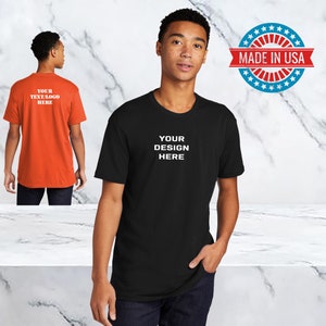 Peut inclure: Un t-shirt noir avec le texte "YOUR DESIGN HERE" en blanc. Le t-shirt est port&eacute; par une personne. L'image comprend &eacute;galement un autocollant en forme d'&eacute;toile rouge, blanc et bleu avec le texte "MADE IN USA".