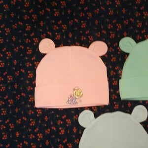 Könnte beinhalten: Drei Baby-Mützen mit Ohrenklappen. Die Mützen sind rosa, hellblau und grün. Jede Mütze hat ein anderes gesticktes Design eines Kindes.