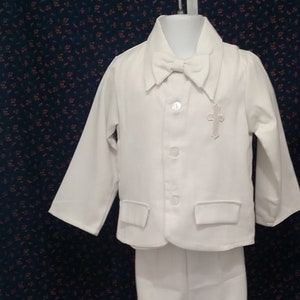 May include: A white suit for a young boy, featuring a jacket with a small cross detail and a bow tie. The suit is hanging on a hanger against a dark blue background.