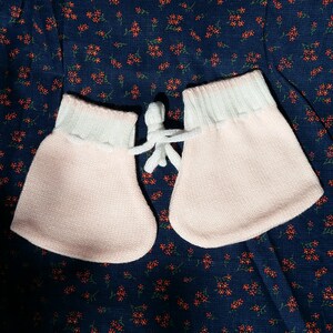 May include: A pair of pink baby mittens with white cuffs. The mittens are on a dark blue fabric with a small red floral pattern.