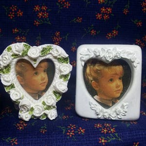 May include: Two white picture frames, one heart-shaped and one rectangular, each holding a portrait of a child. The heart-shaped frame is adorned with white roses and green leaves. The background is a dark blue fabric with small orange floral patterns.