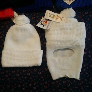 May include: Two white knitted hats with pom-poms. One hat is a traditional beanie style, and the other is a balaclava with a face opening. The hats are on a dark floral patterned background.