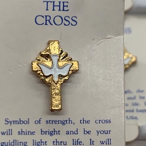Puede incluir: Un broche de cruz dorado con una paloma blanca en el centro. El broche está en una tarjeta con las palabras "THE CROSS" en azul. La tarjeta también incluye texto sobre la cruz como símbolo de fuerza y paz. Hecho en EE. UU.