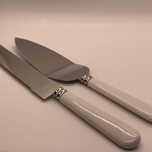 May include: A cake knife and server set. The stainless steel blades have a serrated edge on the knife. Both pieces have white handles with silver-colored accents. The set is designed for serving cake.