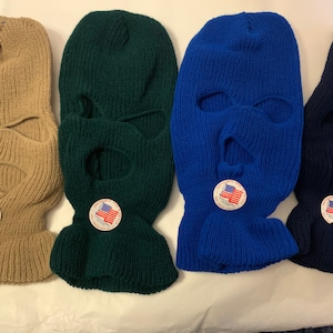 May include: Four knitted balaclavas with eye and mouth holes. The balaclavas are in tan, green, blue, and navy blue. Each balaclava has a small American flag patch.