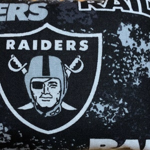 Op de afbeelding: Zwart-witte stof met een herhalend patroon van het Las Vegas Raiders-logo. Het logo toont een schedel met een piratenooglap en gekruiste zwaarden, omringd door het woord "RAIDERS".