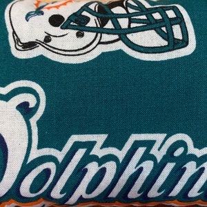 May include: Teal fabric with white stitching and the word "Dolphins" in white with a navy blue outline. A white football helmet with a teal face mask is in the background.