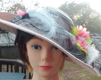 Puede incluir: Un sombrero de ala ancha con un diseño rosa y negro. El sombrero tiene una superposición de malla negra con una cinta rosa y una flor rosa como adorno.