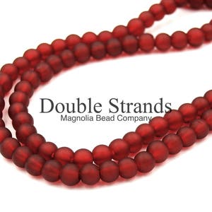May include: Double strands of deep red glass beads. The beads are matte and have a slightly irregular shape. The beads are strung on a clear thread. The image includes the text "Double Strands Magnolia Bead Company".