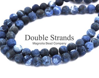 May include: Double strands of blue and white agate beads. The beads are round and have a matte finish. The beads are strung on a clear elastic cord. The image includes the text "Double Strands Magnolia Bead Company".