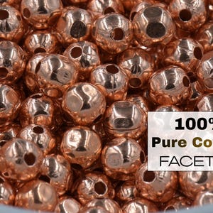 Pode incluir: Close-up de uma pilha de contas de cobre facetadas. As contas são de uma cor cobre brilhante e quente e têm uma forma redonda com múltiplas facetas. O texto "100% Pure Copper FACETED" é visível.