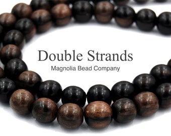 May include: Double strands of dark brown and black wooden beads. The beads are round and have a smooth finish. The beads are arranged in a circle, with the text "Double Strands" and "Magnolia Bead Company" printed below.