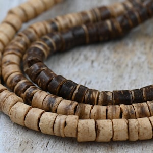May include: Three strands of natural-toned, disc-shaped beads. The beads vary in color from light tan to dark brown, creating a gradient effect. The beads are strung together, forming a necklace or bracelet.