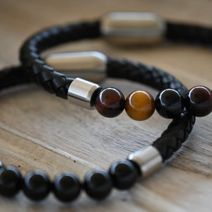 May include: Two black leather braided bracelets with silver clasps. One bracelet features polished, round, black beads. The other has brown and yellow tiger eye beads. The bracelets are a stylish accessory.