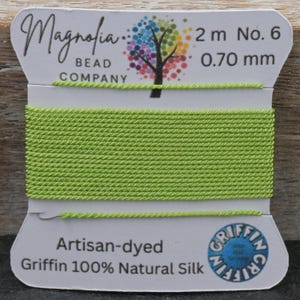 May include: A spool of artisan-dyed, light green Griffin 100% Natural Silk thread. The label reads "Magnolia Bead Company", with details including 2 m length and 0.70 mm thickness. A colorful tree logo is also present.