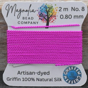 Pode incluir: Um carretel de cordão de seda rosa brilhante enrolado em um cartão branco. O cartão apresenta o texto Magnolia Bead Company, 2 m, No. 8, 0,80 mm, Artisan-dyed, Griffin 100% Natural Silk e um logotipo Griffin. O cordão tem uma textura lisa e torcida.
