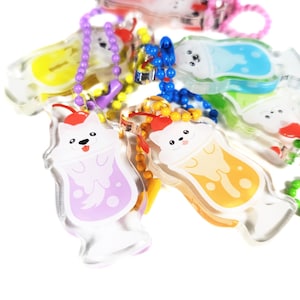 Puede incluir: Una colección de llaveros acrílicos coloridos con diseños de perros de dibujos animados. Cada llavero tiene un esquema de color diferente, incluyendo morado, amarillo, azul y naranja, con una cereza encima. Los llaveros están unidos a cadenas de cuentas.