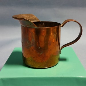 May include: A small copper jug with a hinged lid and a handle. The jug has a worn, aged look and is sitting on a teal surface.