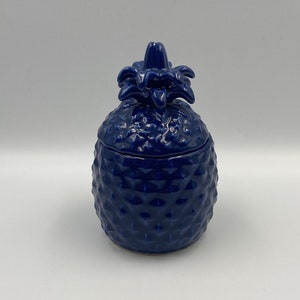 May include: A blue ceramic pineapple shaped jar with a lid. The jar has a textured surface that resembles a pineapple.