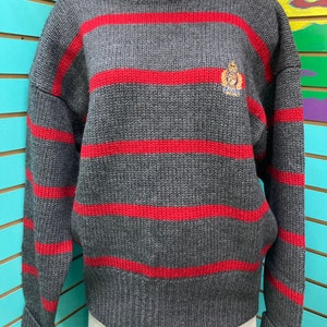 May include: Grey and red striped jumper with a small embroidered logo on the chest. The logo features a crown and a horse.