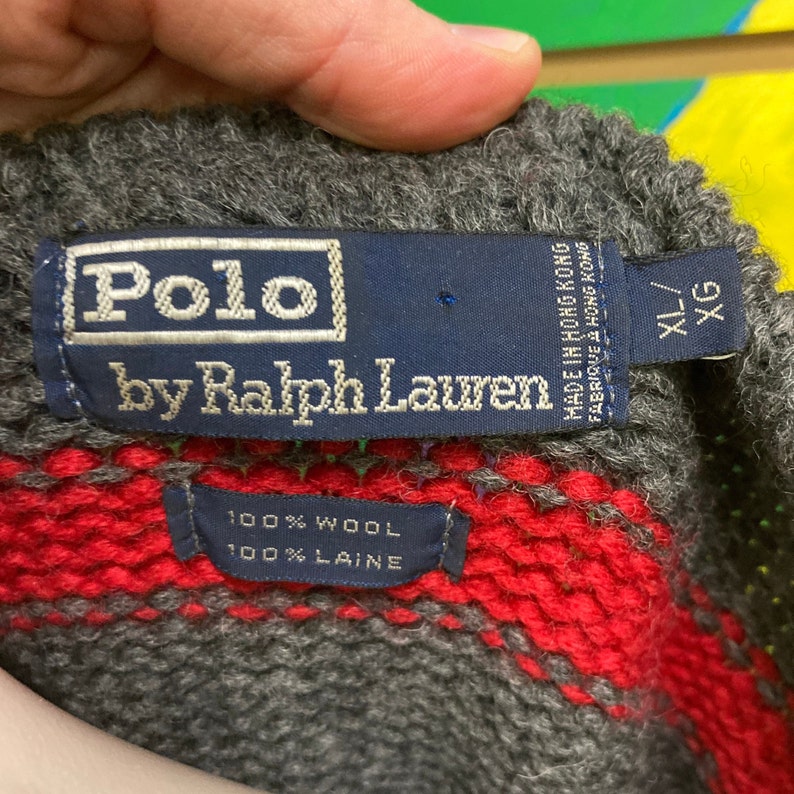 May include: A grey and red striped wool jumper with a blue Polo by Ralph Lauren label. The label says "Made in Hong Kong" and "100% Wool/100% Laine". The jumper has a size tag that says "XL/XG".