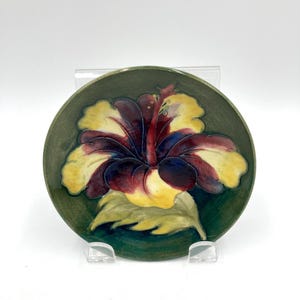 May include: A circular ceramic plate featuring a hand-painted hibiscus flower design. The flower has yellow, burgundy, and blue petals with a red centre, set against a green background. The plate is displayed on a clear stand.