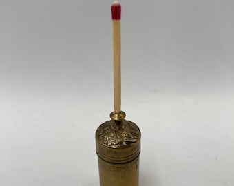 May include: A small, ornate brass container with a matchstick inserted into the top. The container has a decorative floral design and a lid with a knob.