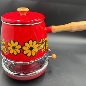 May include: A vintage red enamel pot with a wooden handle and lid knob. The pot features yellow and black daisy floral designs. It sits on a chrome stand with a small wooden knob. A retro kitchenware item.