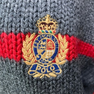 May include: A grey knitted sweater with a red and white stripe and a large embroidered crest featuring a crown, shield, and the word "POLO".