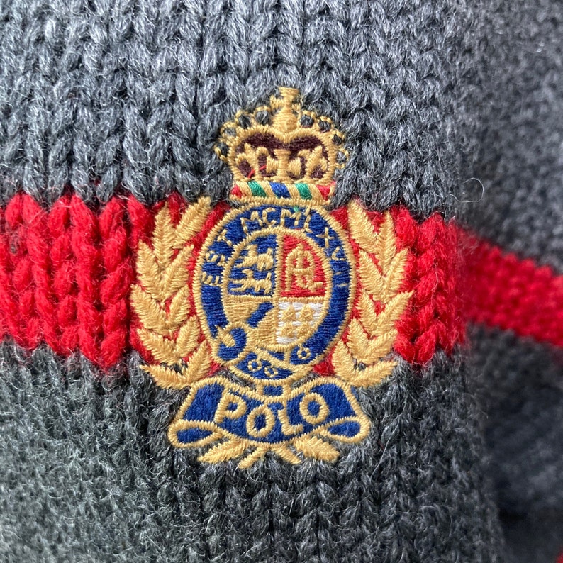 May include: A grey knitted sweater with a red and white stripe and a large embroidered crest featuring a crown, shield, and the word "POLO".