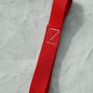 Puede incluir: Un lazo de cinta roja con costuras blancas. El lazo está diseñado para ser utilizado como asa o punto de fijación.