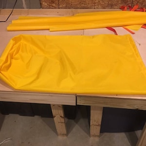 Puede incluir: Una tela de color amarillo brillante, posiblemente nailon, está extendida sobre una superficie de trabajo de madera. La tela está doblada y arrugada, con un cordón visible. También hay piezas de tela amarilla adicionales y tiras naranjas.