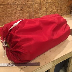 Può includere: Un sacco a pelo rosso con una striscia bianca. Il sacco è arrotolato e legato con uno spago bianco.