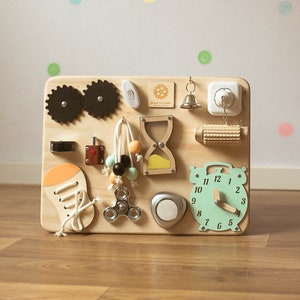 May include: A wooden activity board for toddlers featuring various interactive elements. Includes gears, a light switch, a bell, a plug, a roller, a clock, a spinner, an hourglass, a lock, and a shoe with laces. The board is made of light-colored wood.