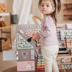 May include: A young girl plays with a wooden activity board with various textures and activities. The board is labeled "VIOLET" and features a colorful xylophone, gears, a door, a lock, and a maze. The girl is wearing a light purple long-sleeved shirt and white pants.