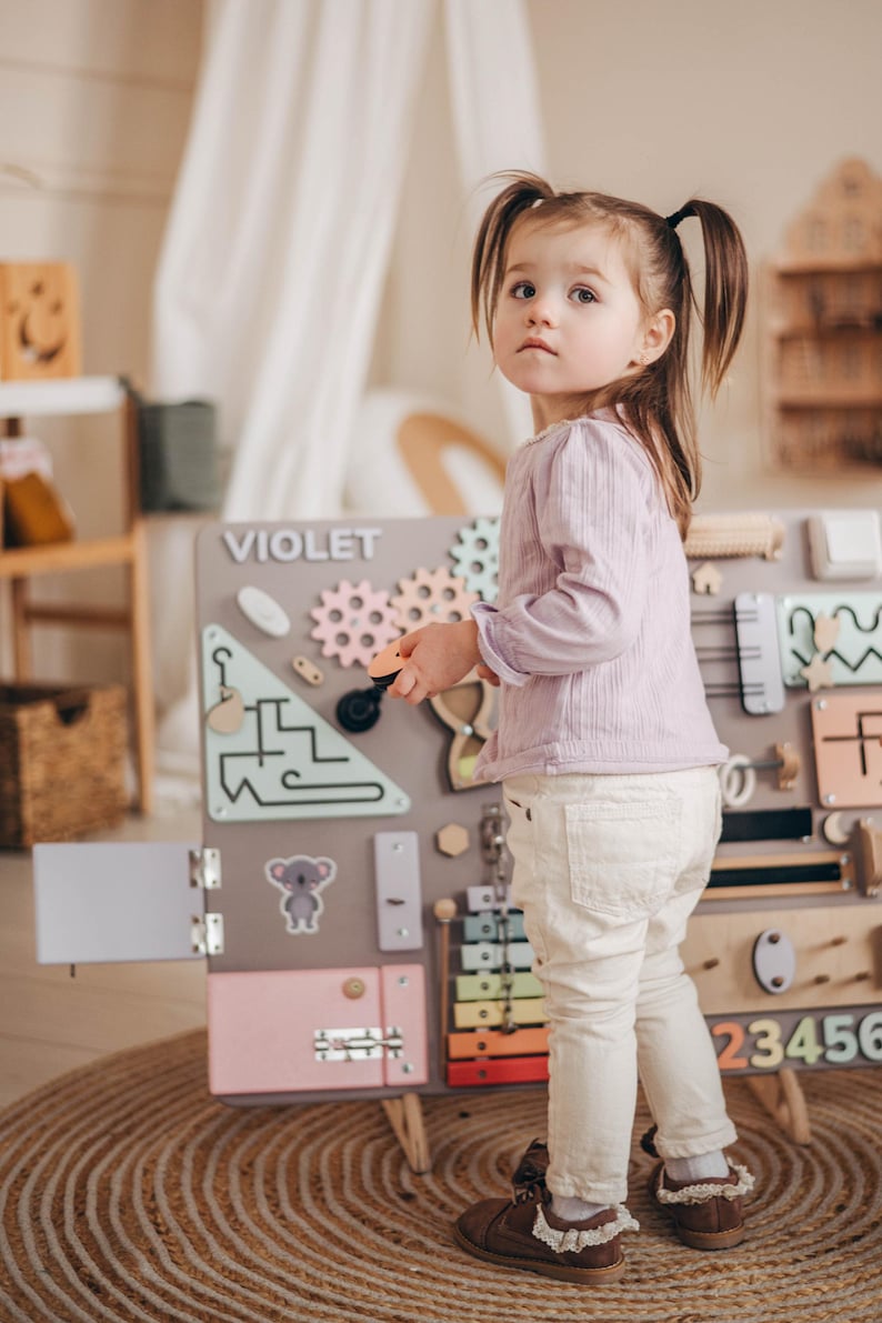 May include: A young girl plays with a wooden activity board with various textures and activities. The board is labeled "VIOLET" and features a colorful xylophone, gears, a door, a lock, and a maze. The girl is wearing a light purple long-sleeved shirt and white pants.
