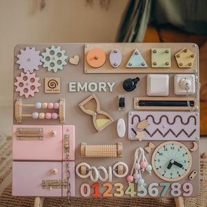 May include: A wooden activity board with various interactive elements for children. Features include gears, shapes, a clock, a drawer, and the name "EMORY". The board is primarily pastel colors with wooden accents, and includes the numbers 0-9.