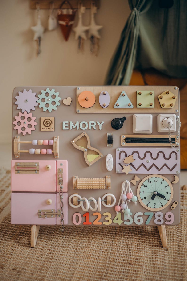 May include: A wooden activity board with various interactive elements for children. Features include gears, shapes, a clock, a drawer, and the name "EMORY". The board is primarily pastel colors with wooden accents, and includes the numbers 0-9.