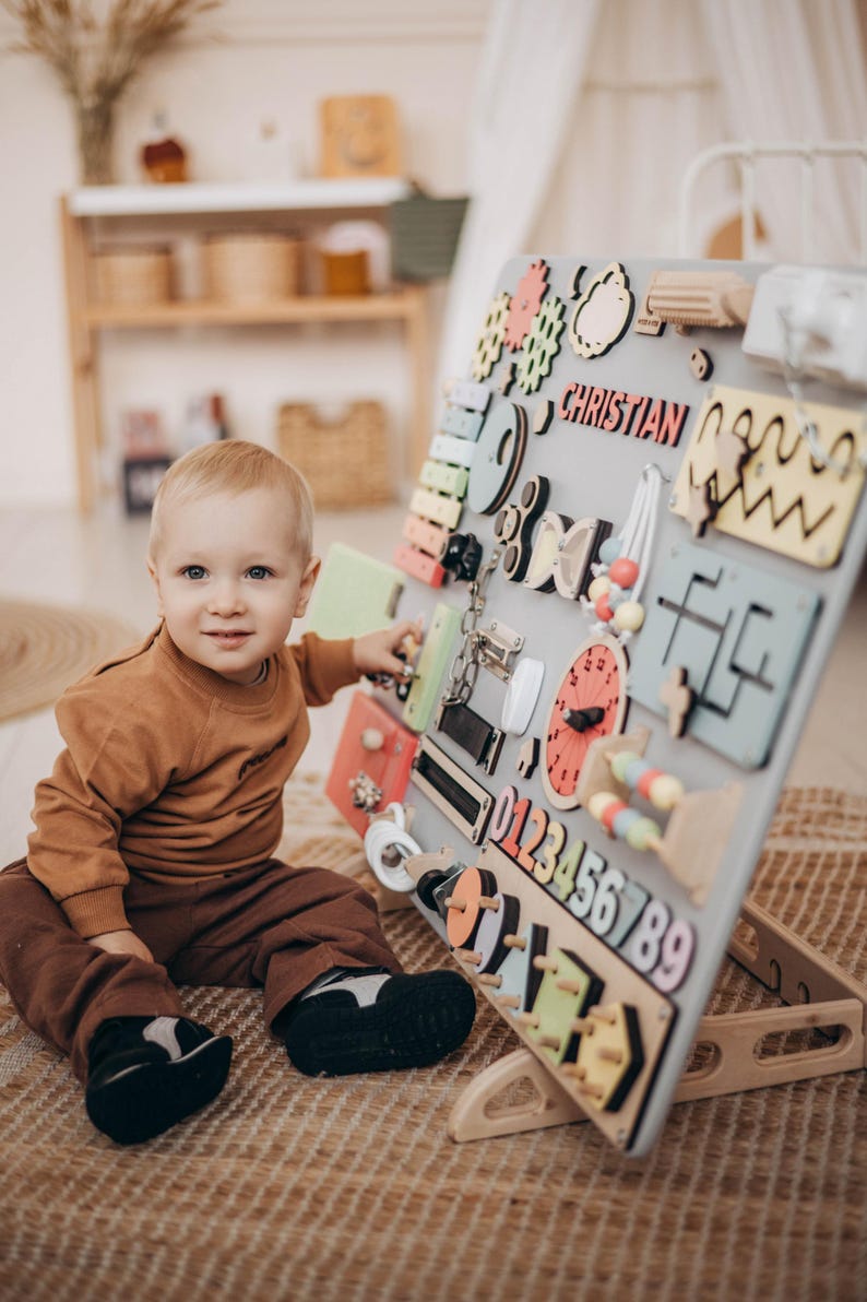 May include: A toddler plays with a colorful wooden activity board. The board has various textures, shapes, and activities, including a clock, a lock, a slide, and numbers. The board is personalized with the name "Christian".