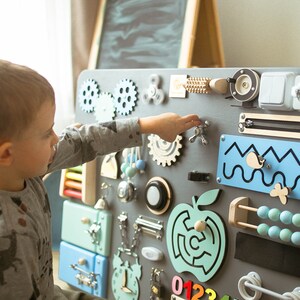 May include: A gray wooden busy board with various interactive elements for children. Features include gears, a spinner, a zipper, a light switch, an abacus, and a maze. The board is designed to stimulate fine motor skills and sensory exploration.
