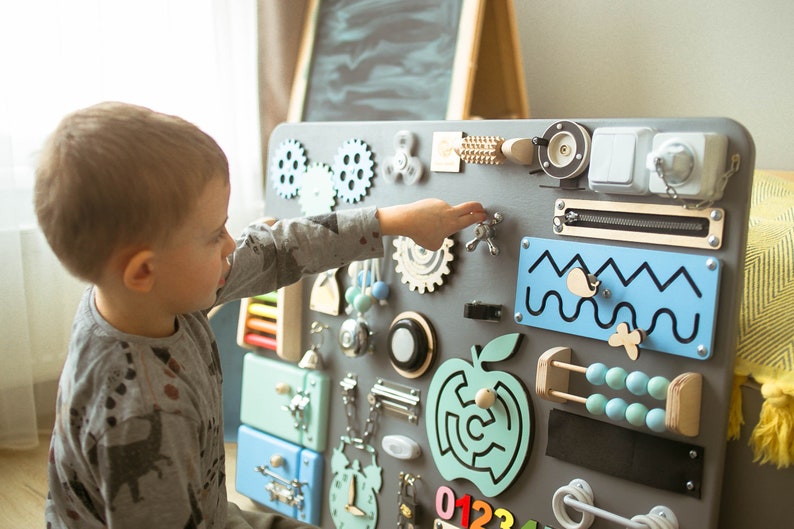 May include: A gray wooden busy board with various interactive elements for children. Features include gears, a spinner, a zipper, a light switch, an abacus, and a maze. The board is designed to stimulate fine motor skills and sensory exploration.