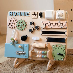 May include: A wooden activity board with a variety of textures and activities for children. The board is painted in natural wood tones and features a blue latch, a clock, a spinning fidget, gears, a bead maze, and a slider. The board is personalized with the name "BONNIE" and the logo "WOOD & KIDS".