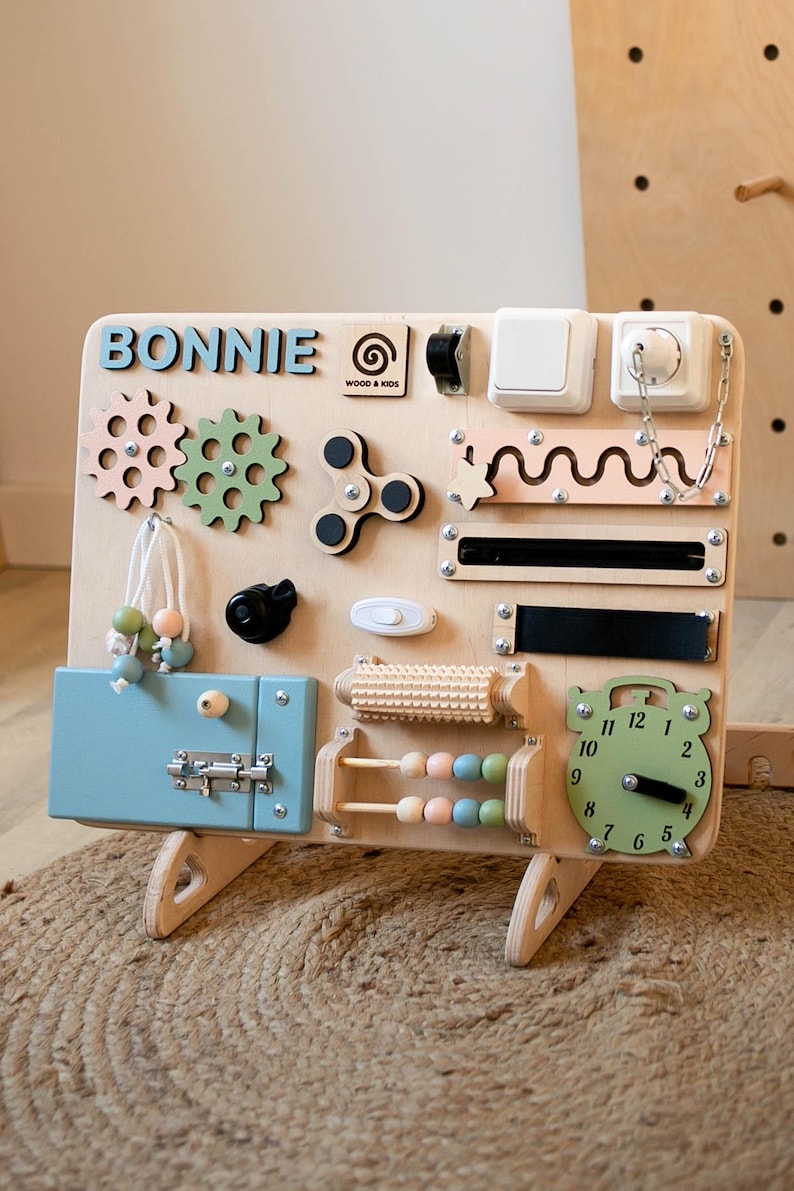 May include: A wooden activity board with a variety of textures and activities for children. The board is painted in natural wood tones and features a blue latch, a clock, a spinning fidget, gears, a bead maze, and a slider. The board is personalized with the name "BONNIE" and the logo "WOOD & KIDS".