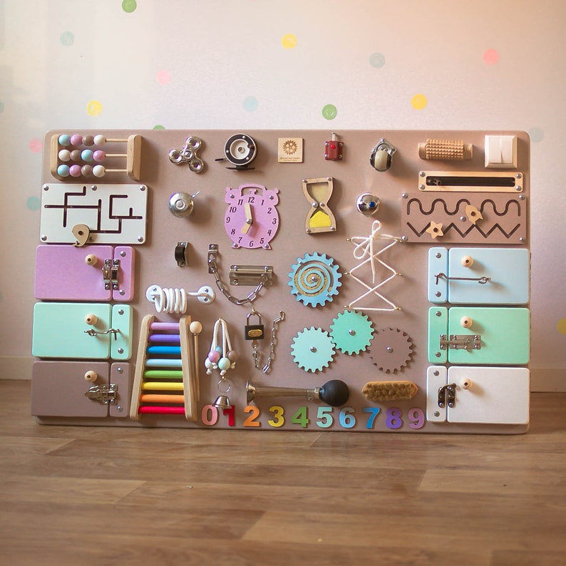May include: A large, wooden busy board with various interactive elements for children. Features include colorful drawers, locks, gears, an abacus, a clock, and a xylophone. The board is primarily beige with pastel-colored accents and wooden components.