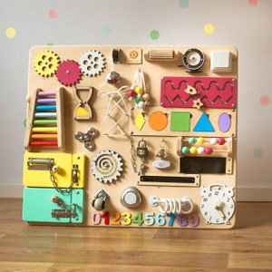 May include: A wooden activity board with various interactive elements for children. Features include gears, a colorful abacus, a clock, latches, a spinner, and a small ladder. The board has a natural wood finish with colorful accents and shapes.