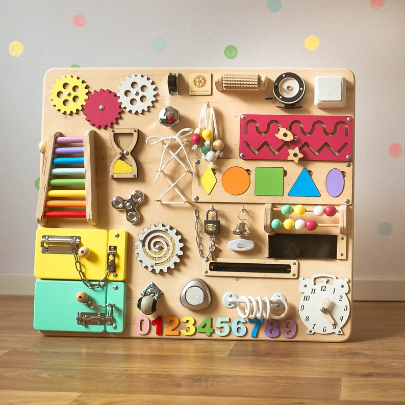 May include: A wooden activity board with various interactive elements for children. Features include gears, a colorful abacus, a clock, latches, a spinner, and a small ladder. The board has a natural wood finish with colorful accents and shapes.